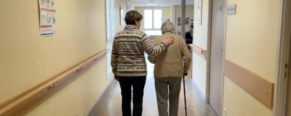 Une fille et sa mère âgée marchent ensemble dans un couloir d'EHPAD lumineux lors d'une visite de pré-admission